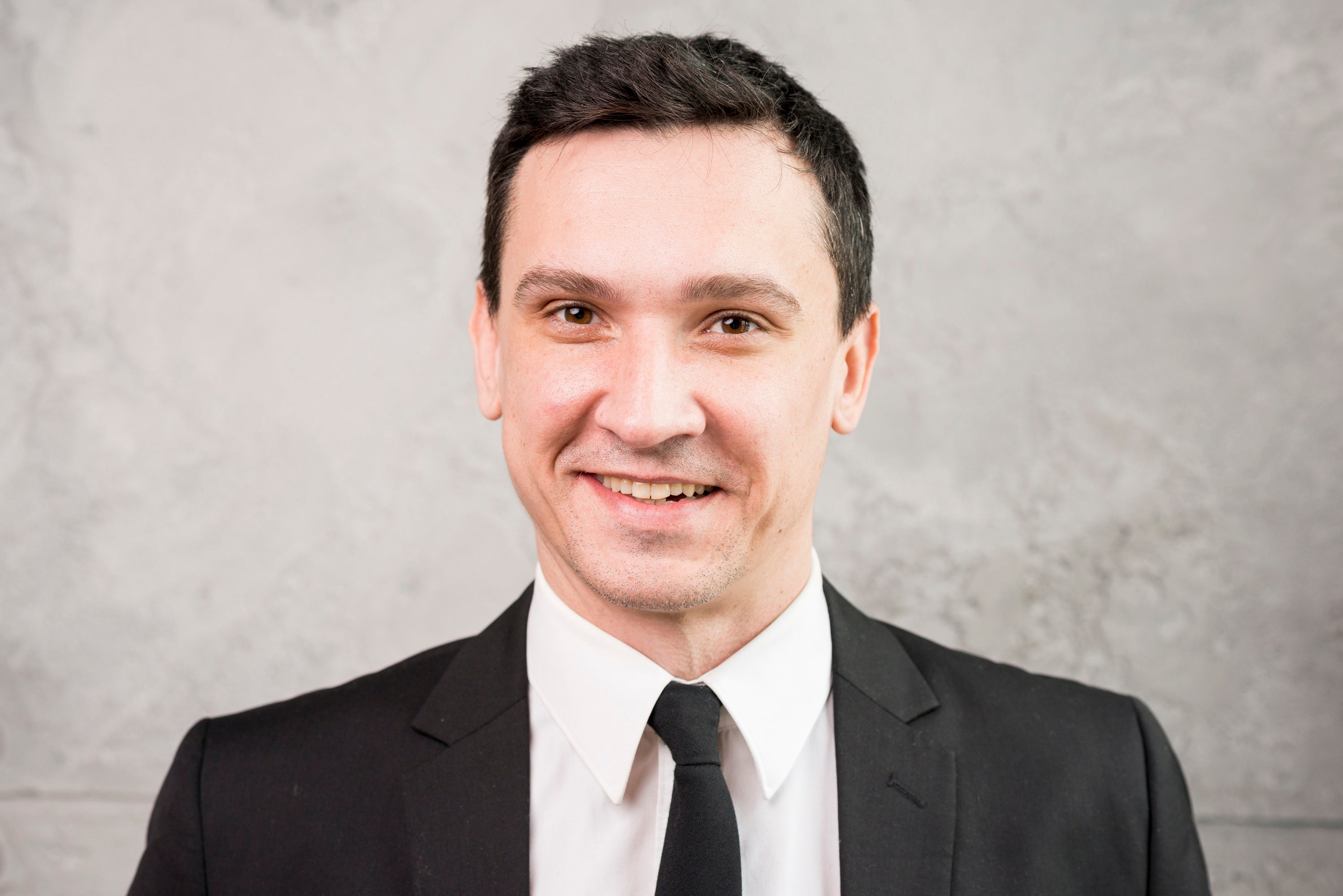 Man wearing a black suit and tie against a gray background